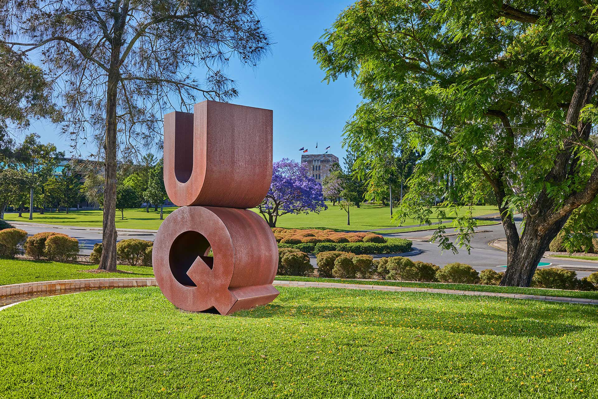 bronze sign with the letters UQ on a grass field with sandstone buildings and trees in the background