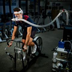 A man cycling on a gym machine, while his breathing is measured via a breathing tube.