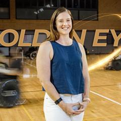 Emma Beckman on a basketball court with the words "golden eye" behind her as well ass blurry wheelchair basketballers.